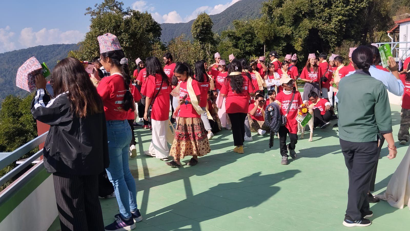 Campers in red shirts and guests in Nepali dress gathered on a terrace above the valley