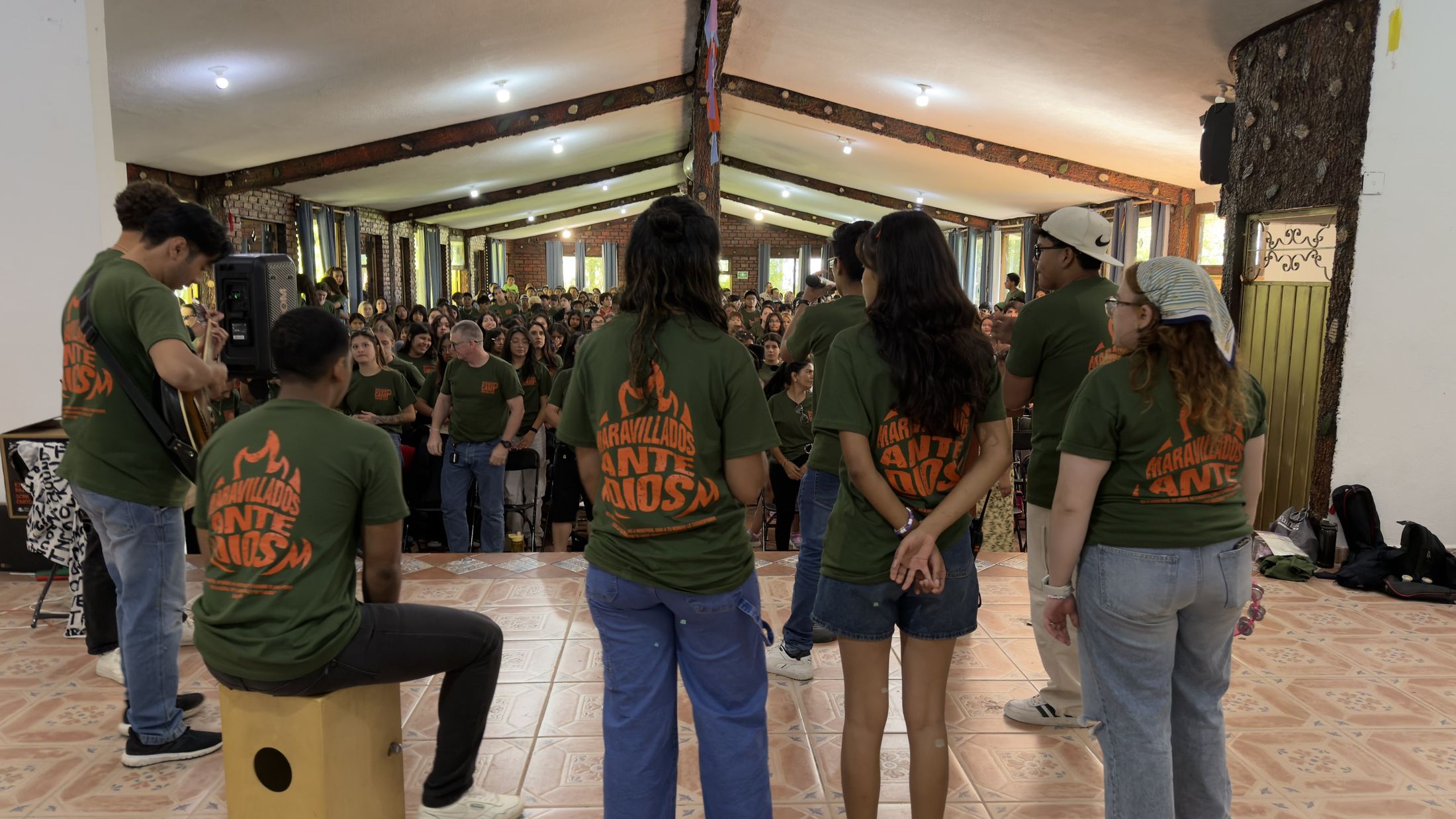 Counselors in green camp shirts, seen from behind, facing a full dining hall of campers