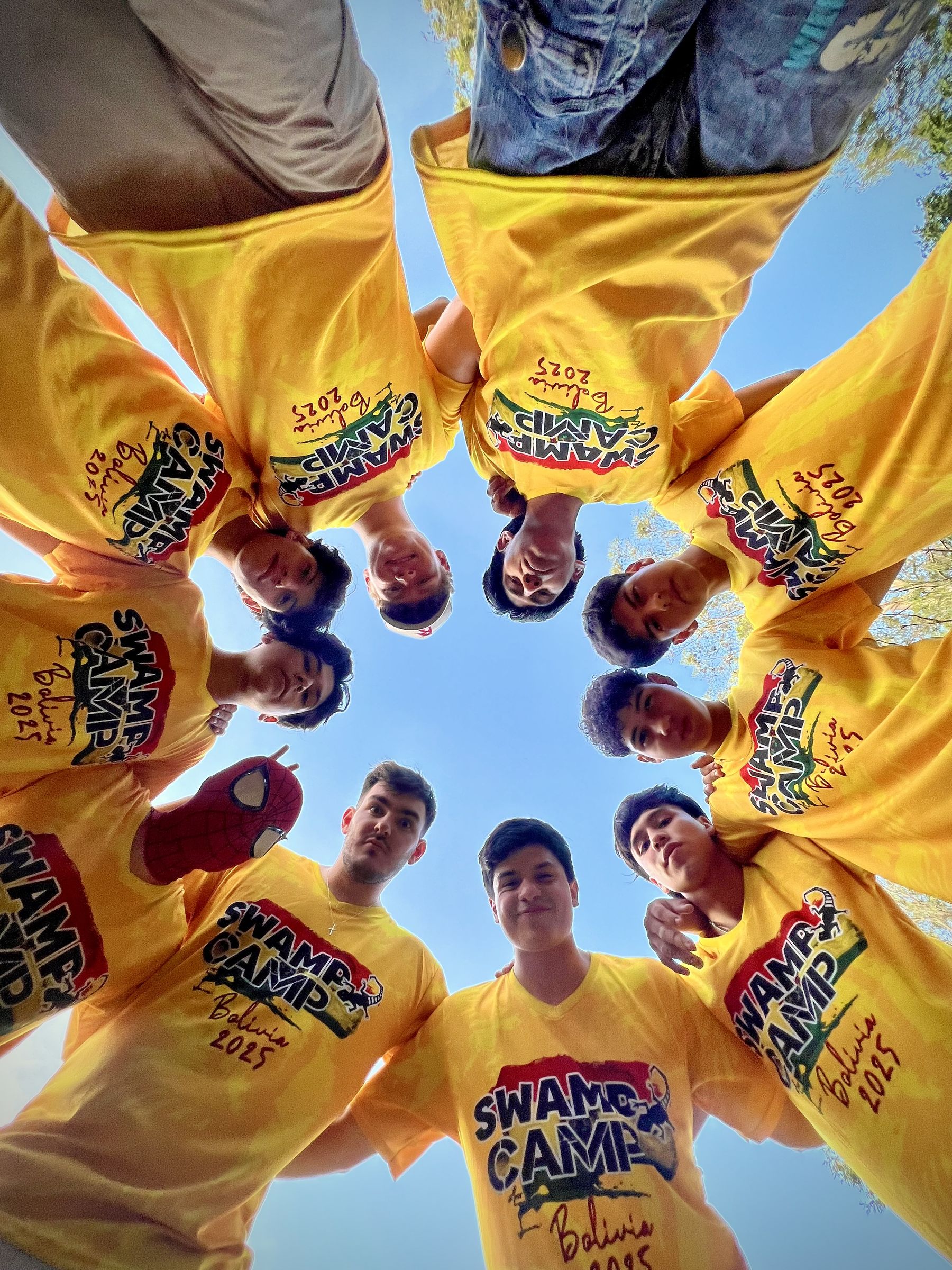 A team in yellow Swamp Camp shirts huddled in a circle, seen from below against the sky