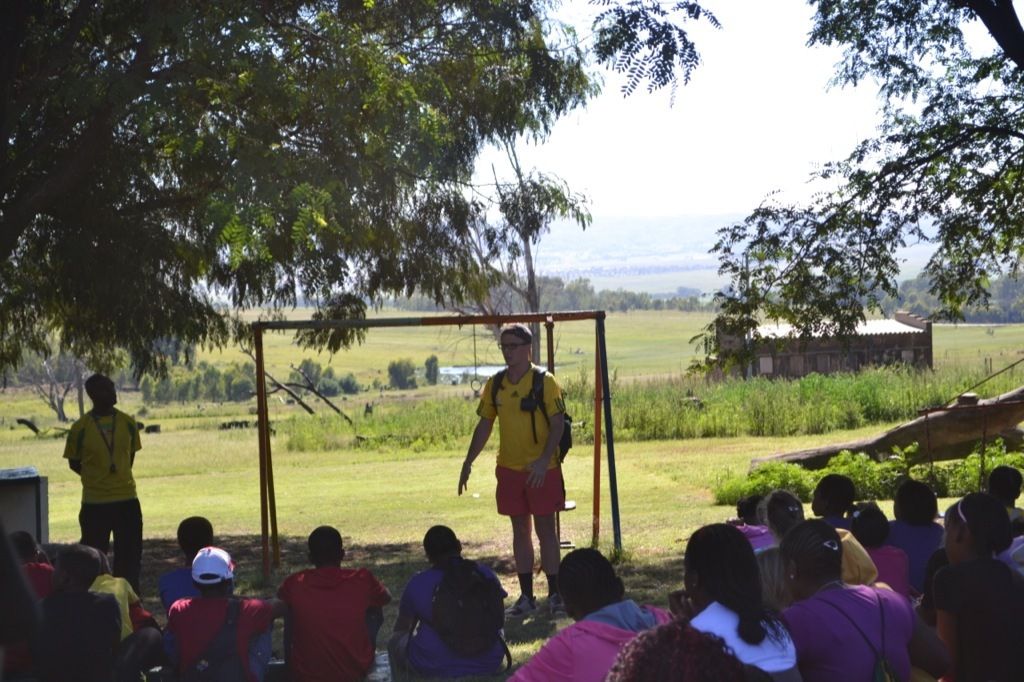 A boy standing to speak in front of the whole camp seated under the trees