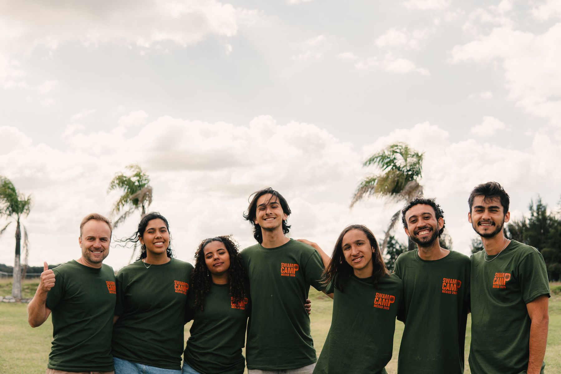 Seven counselors in green Swamp Camp Mexico shirts, arms around each other, palms and sky behind